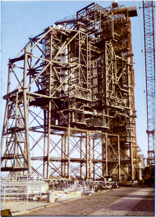 Image 007. Rising nearly 200 feet above the level of the Crawlerway you’re looking at it from (which itself rises nearly 50 feet above the level of the surrounding wetlands), the Rotating Service Structure at Space Shuttle Launch Complex 39-B, Kennedy Space Center, Florida, stands as an open skeleton, still partially supported on the falsework which the Union Ironworkers working for Wilhoit Steel Erectors constructed first, to hold it up temporarily while it was being erected, before it became self-supporting and the falsework could then be removed. Behind the gray steel of the RSS, the red framework of the taller Fixed Service Structure, which was built first, reusing parts of an Apollo Project Launch Umbilical Tower, can also be seen. On the pad deck in the foreground beneath the falsework, additional material fabricated by Sheffield Steel, who fabricated all of the structural steel for the RSS, can be seen, awaiting its turn to be lifted into place by Wilhoit’s big Manitowoc crane, visible along the right margin of the image. Photo by James MacLaren. Image 007. Rising nearly 200 feet above the level of the Crawlerway you’re looking at it from (which itself rises nearly 50 feet above the level of the surrounding wetlands), the Rotating Service Structure at Space Shuttle Launch Complex 39-B, Kennedy Space Center, Florida, stands as an open skeleton, still partially supported on the falsework which the Union Ironworkers working for Wilhoit Steel Erectors constructed first, to hold it up temporarily while it was being erected, before it became self-supporting and the falsework could then be removed. Behind the gray steel of the RSS, the red framework of the taller Fixed Service Structure, which was built first, reusing parts of an Apollo Project Launch Umbilical Tower, can also be seen. On the pad deck in the foreground beneath the falsework, additional material fabricated by Sheffield Steel, who fabricated all of the structural steel for the RSS, can be seen, awaiting its turn to be lifted into place by Wilhoit’s big Manitowoc crane, visible along the right margin of the image. Photo by James MacLaren.